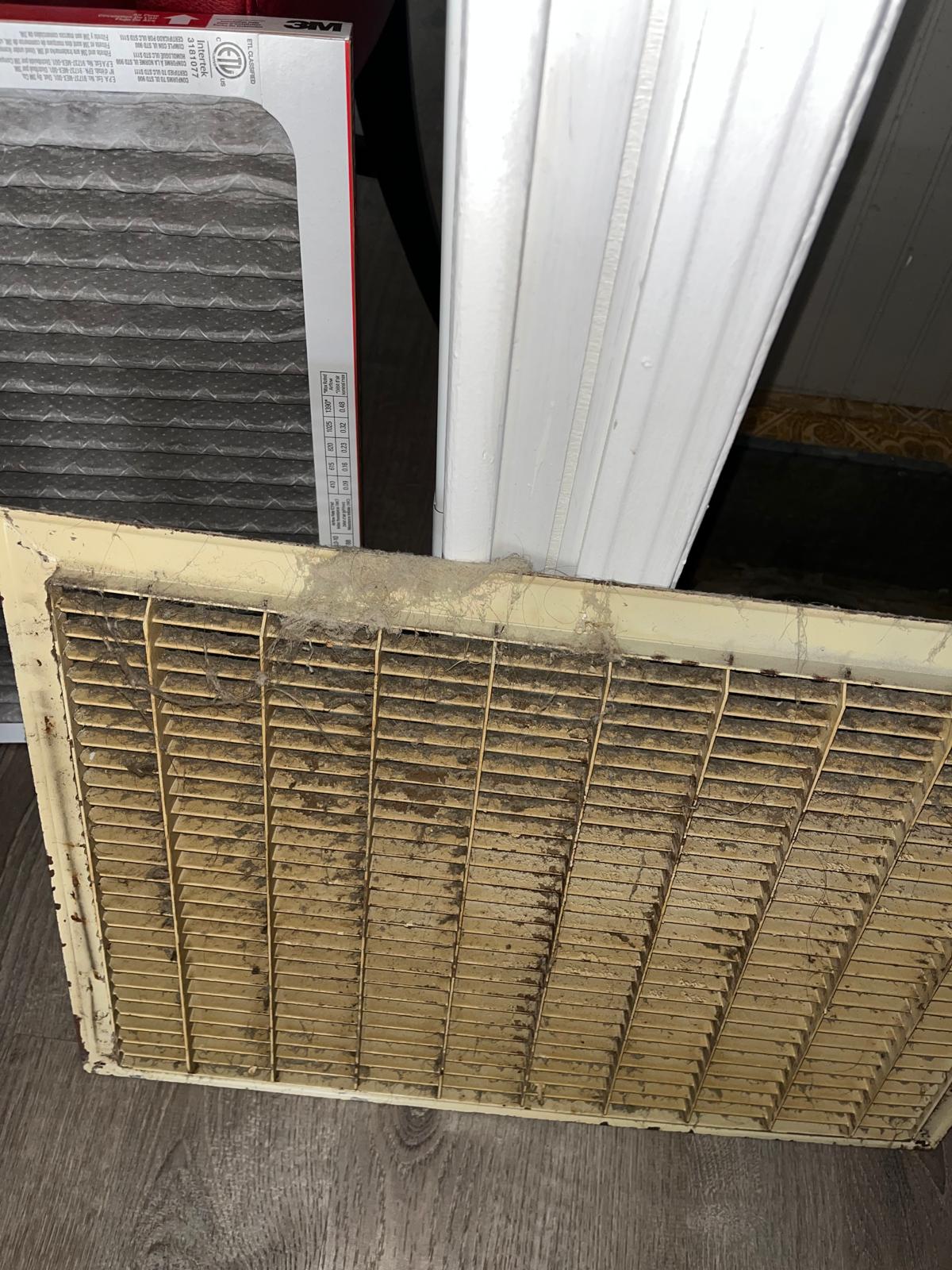 Wide-angle view of a ceiling-mounted air return vent heavily coated with dust buildup beside its replacement filter